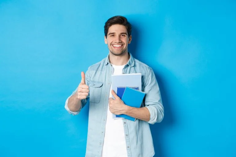 Young Man Student with Notebooks Showing Thumb Up Approval Smiling Satisfied Blue Studio Background - YES Germany